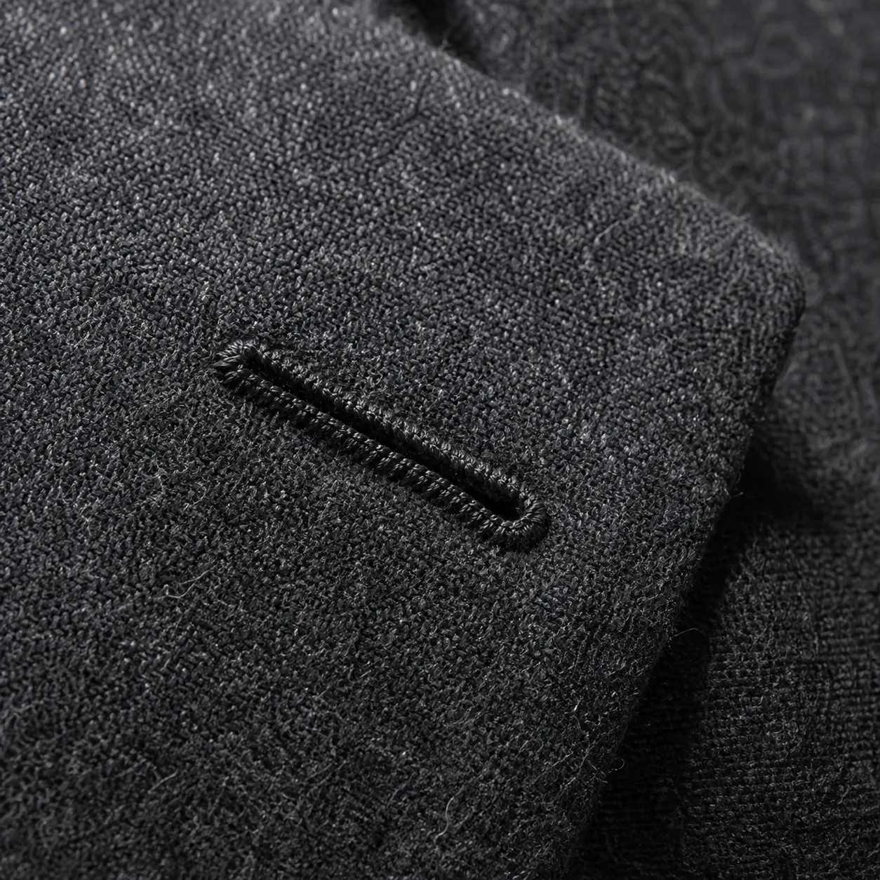 Extreme close-up of a hand-stitched Milanese buttonhole on a charcoal peak lapel, thread catching raking light against the wool nap.